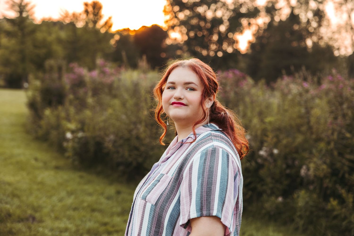 A high school senior wearing a striped button-up shirt poses in a garden at sunset with golden lighting. Metro Detroit Senior Photography