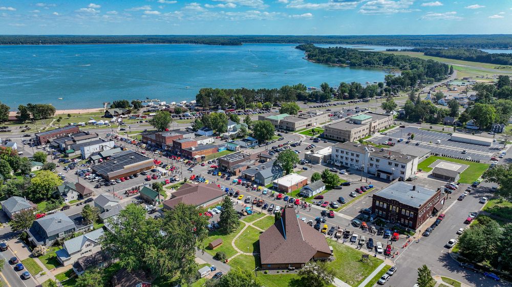 Aerial view of a small lakeside town with colorful classic cars parked along the main street during a car show.