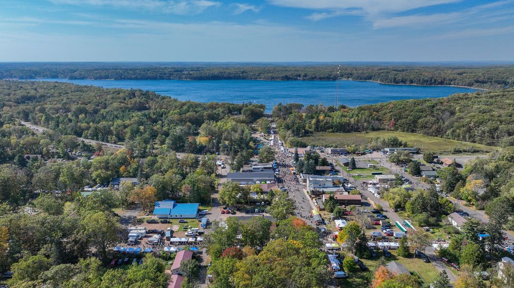 Bird's-eye view of a charming small town with a vibrant main street, colorful trees, and a large blue lake behind it.