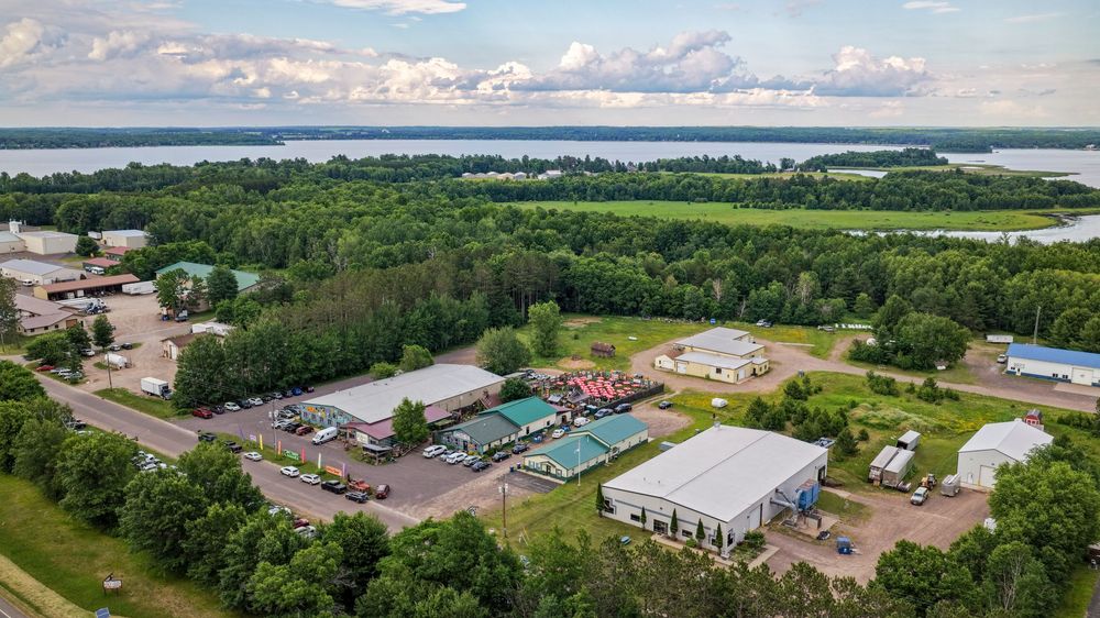 Aerial view of a rural commercial property with green roofed buildings, parking lot, and a river with tree-lined shores in background.