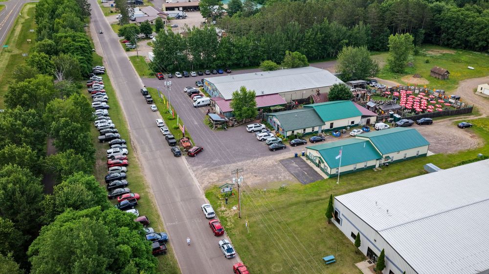 Aerial view of a roadside commercial property with green roofed building, large parking lot filled with cars, and surrounding greenery.