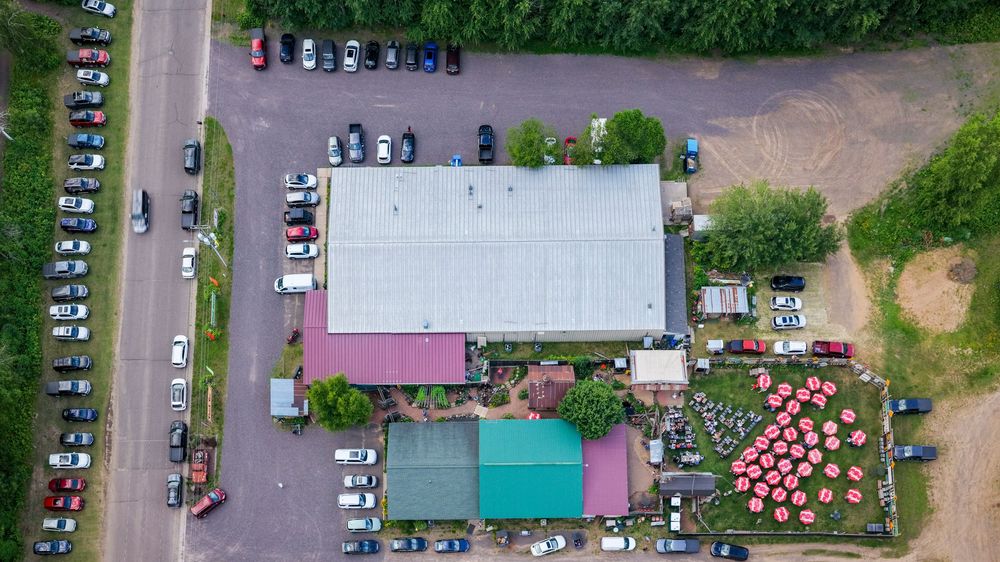 Aerial view of a rural event venue with large warehouse, parking lot, and colorful outdoor seating area surrounded by trees.