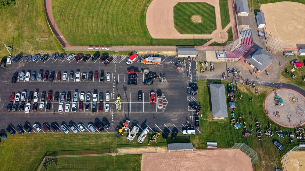 Aerial view of a packed parking lot beside a baseball field and recreational facilities on a sunny day.
