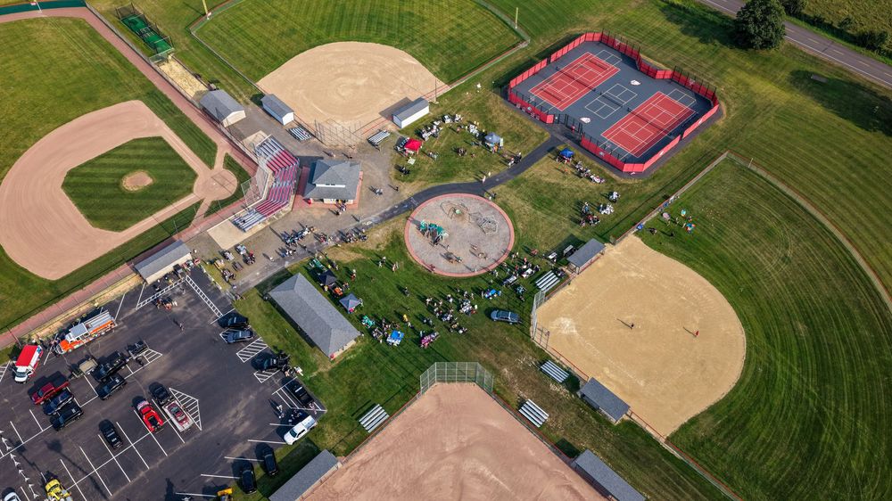 Aerial view of a sports complex featuring baseball fields, tennis courts, and a parking lot surrounded by green farmland.