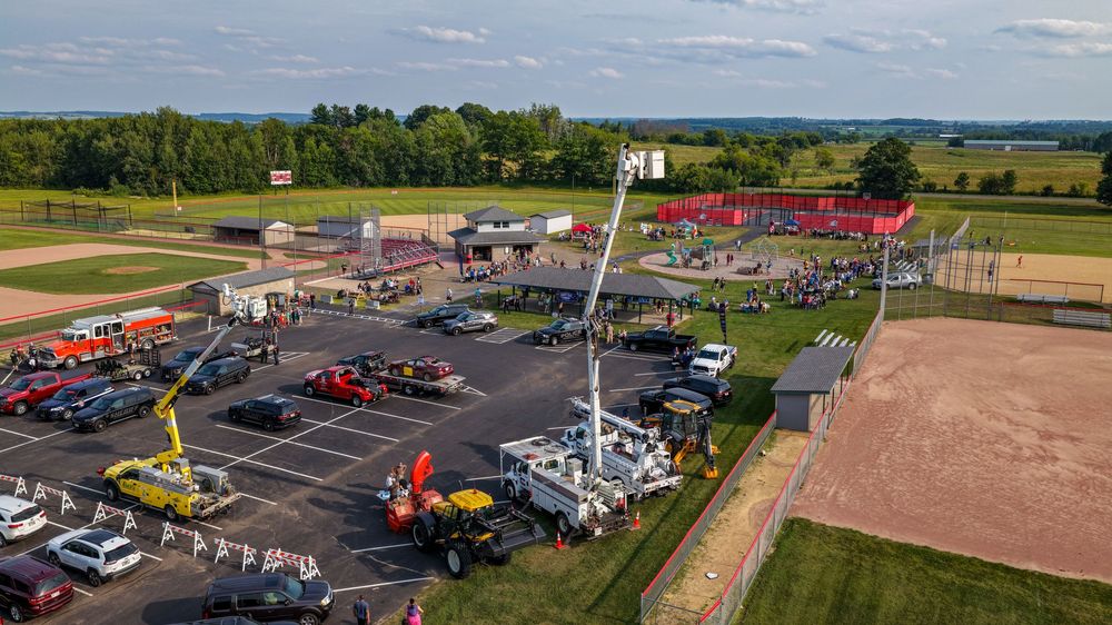 Aerial view of a busy parking lot with cranes, emergency vehicles, and crowds near a red building surrounded by green fields.