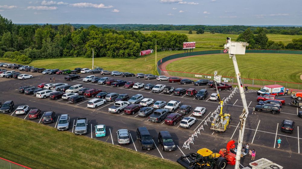 Aerial view of a packed parking lot near green fields, with a boom lift and red building visible in the background.