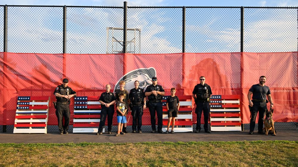 Police officers and K9 unit pose with children in front of red fence with American flag displays outdoors.