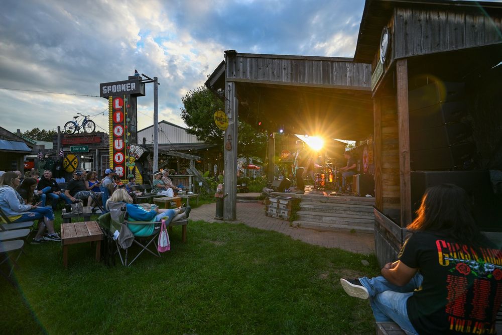 Outdoor concert at sunset near a rustic saloon with crowd enjoying live music on a lush green lawn.