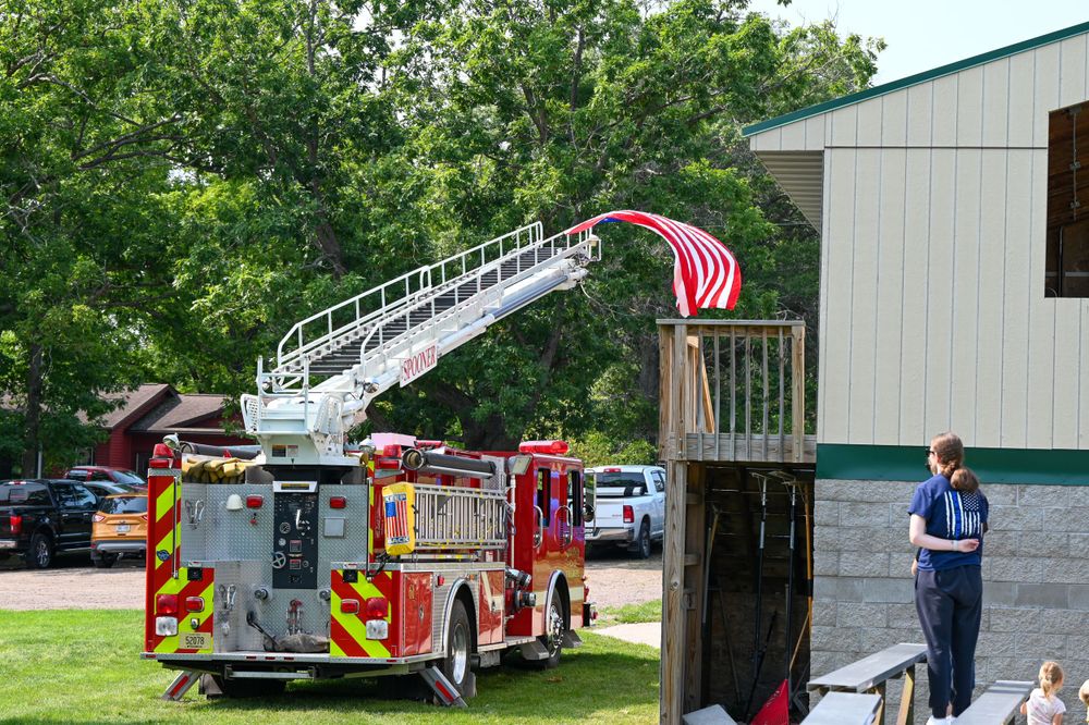 Red fire truck with ladder holding American flag beside a building with firefighters nearby.