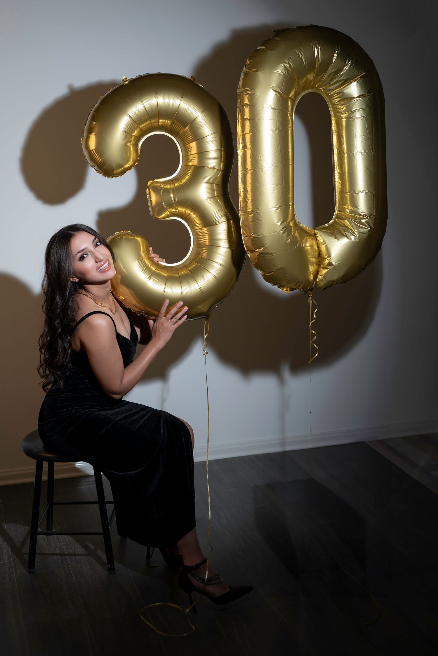 Gold metallic number balloons showing '30' against a dark wall with someone in a black dress seated on a stool.