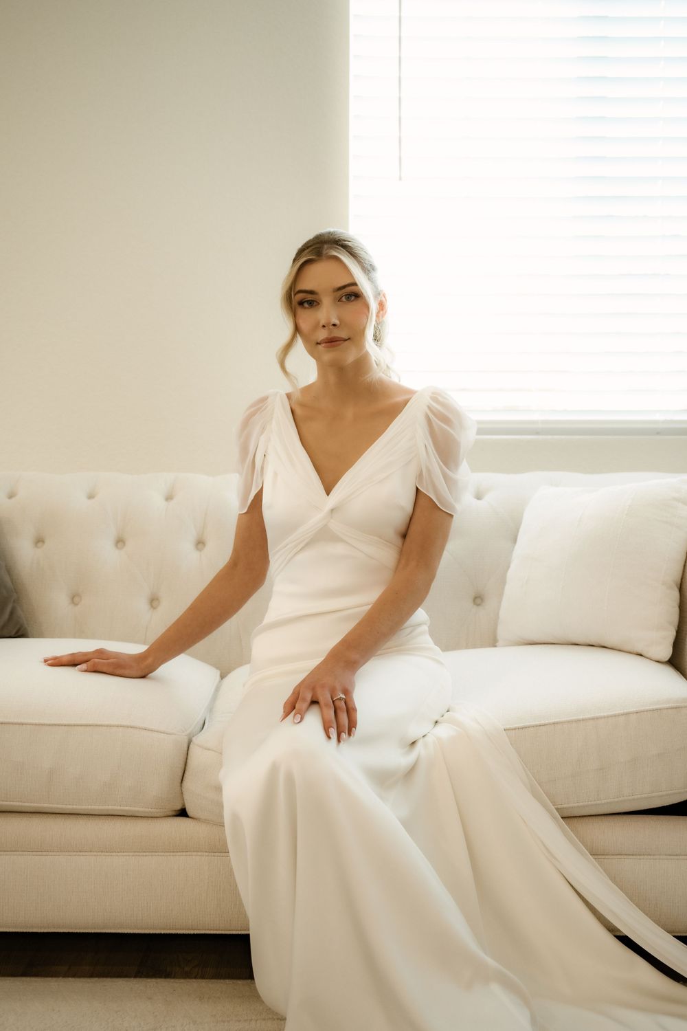 Elegant white wedding dress displayed on a cream tufted sofa in natural window light.