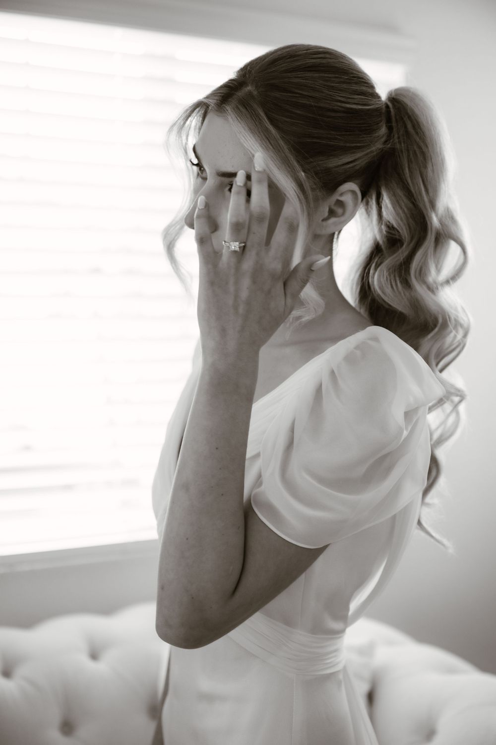 Emotional bridal moment captured in black and white as hands cover face while seated on white sofa.