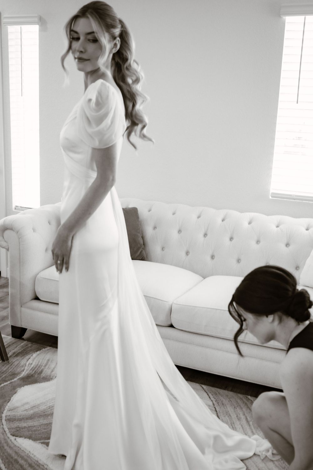 Elegant white wedding dress being fitted in a bright room with a tufted sofa.