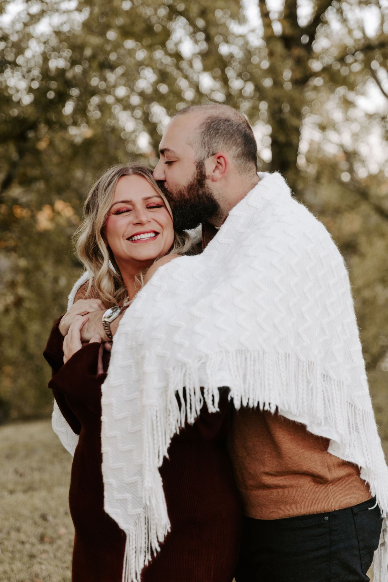 Romantic couple sharing intimate moments outdoors while wrapped in a white fringed blanket against an autumn backdrop.