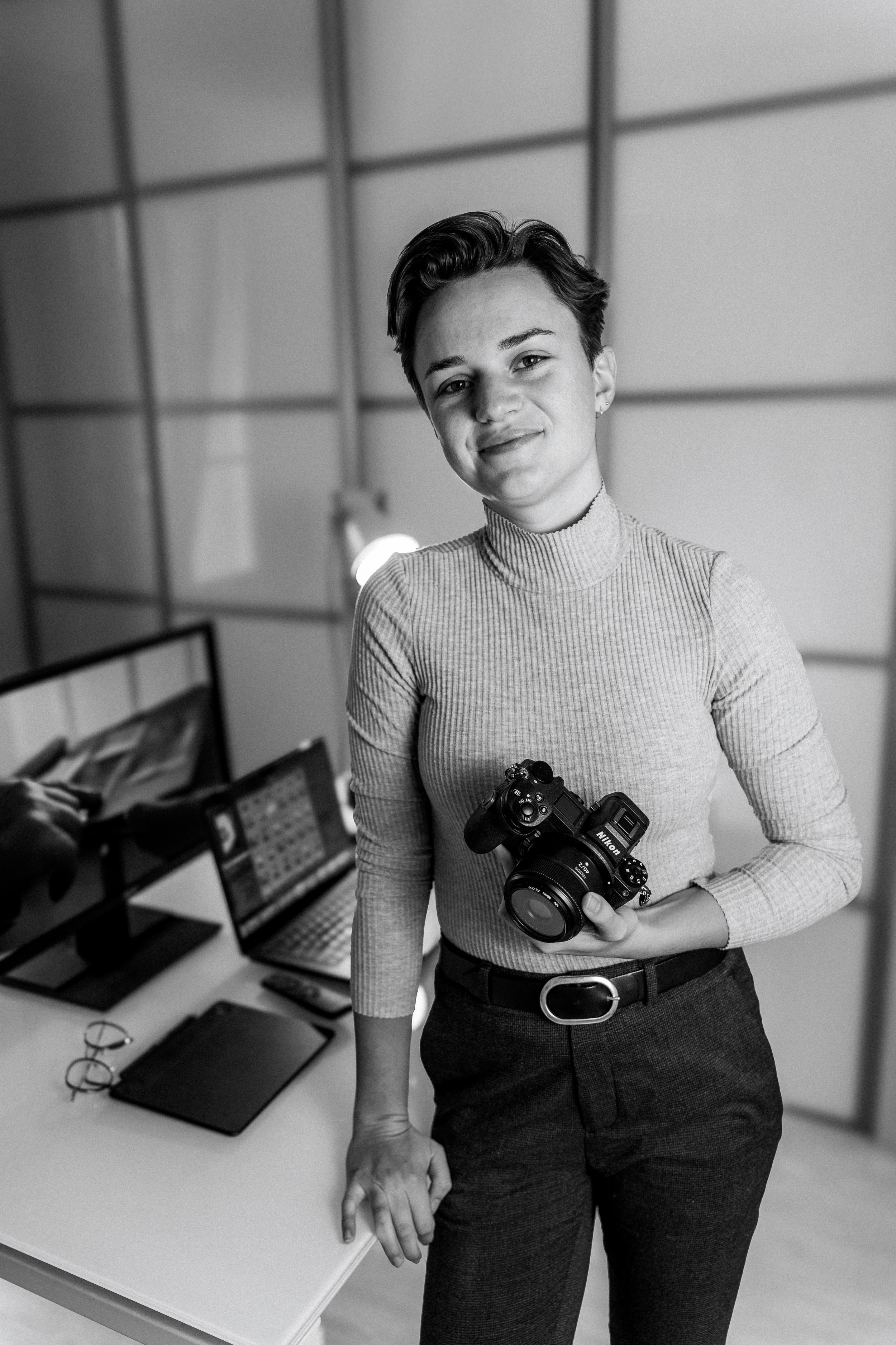 A photographer poses with a camera in an office setting wearing a grey turtleneck and dark pants.