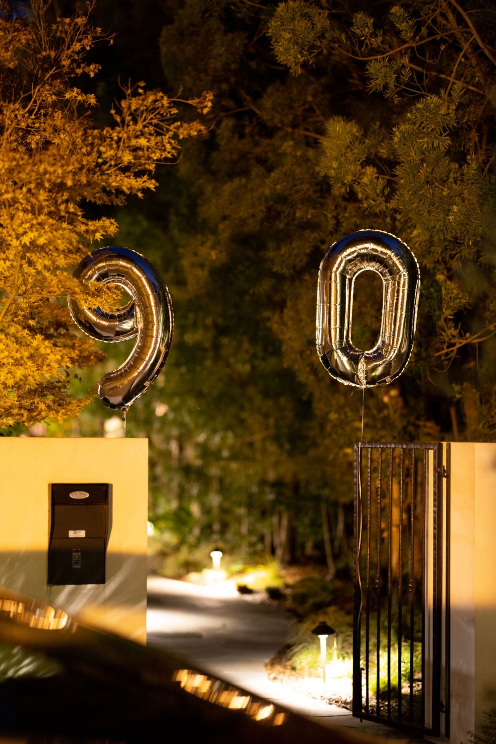 Gold number 90 balloons float above a gated driveway entrance at night surrounded by illuminated trees.