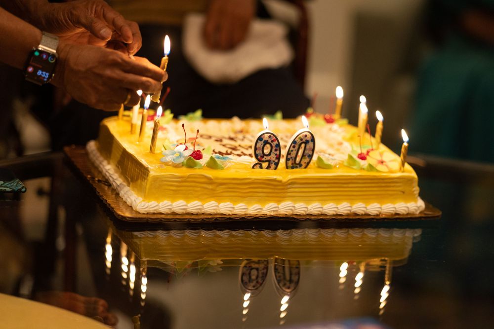 Hands lighting candles on a 90th birthday cake decorated with number 90 candles on a reflective surface.