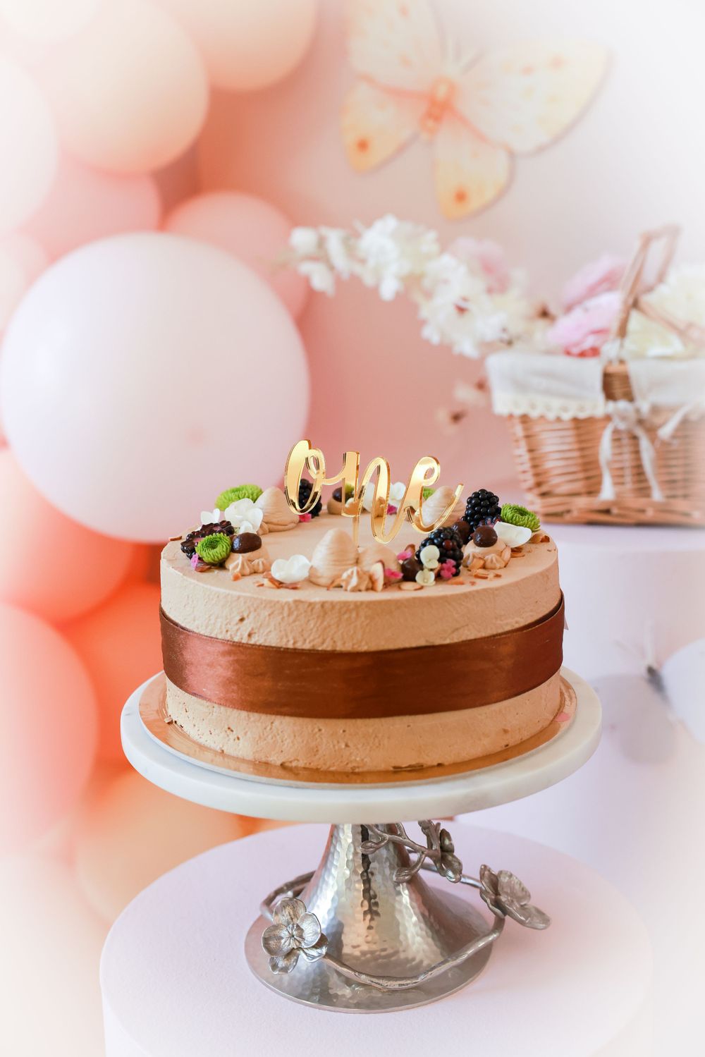 Elegant first birthday cake on silver stand topped with gold one topper and fresh berries among pink balloon decor.