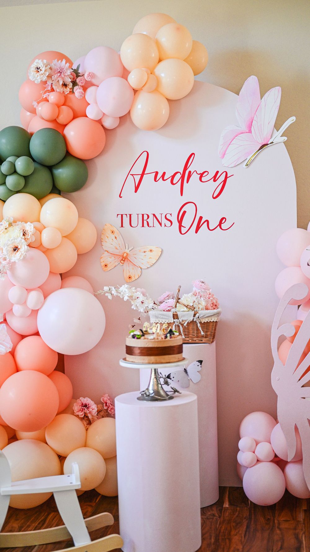 First birthday party backdrop reading Audrey Turns One with pink and peach balloon garland and butterfly decorations.