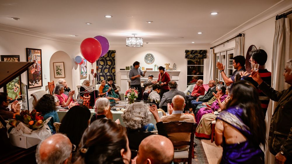 Large group of people celebrating at an indoor 90th birthday party with colorful balloons in a warmly lit home.
