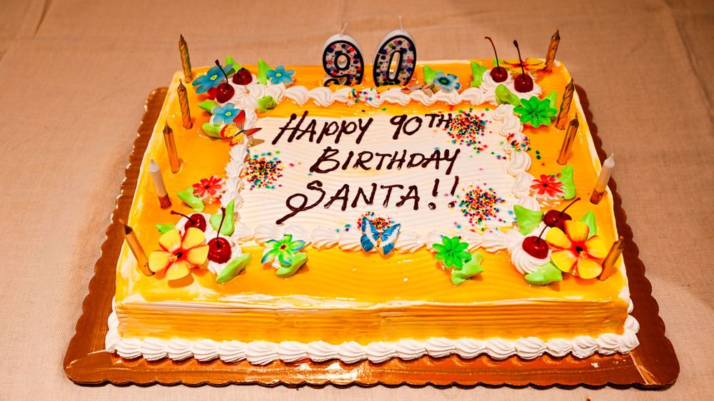 Rectangular birthday cake with orange frosting reads Happy 90th Birthday Santa decorated with tropical flowers.