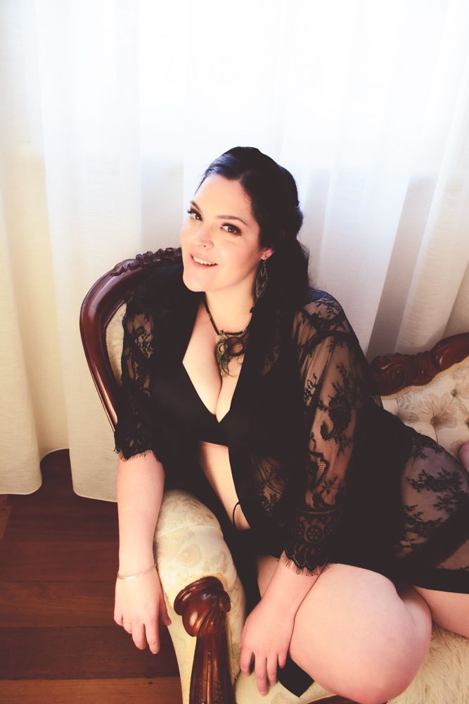 A boudoir photo featuring a black lace robe and vintage chair against light curtains and hardwood flooring.