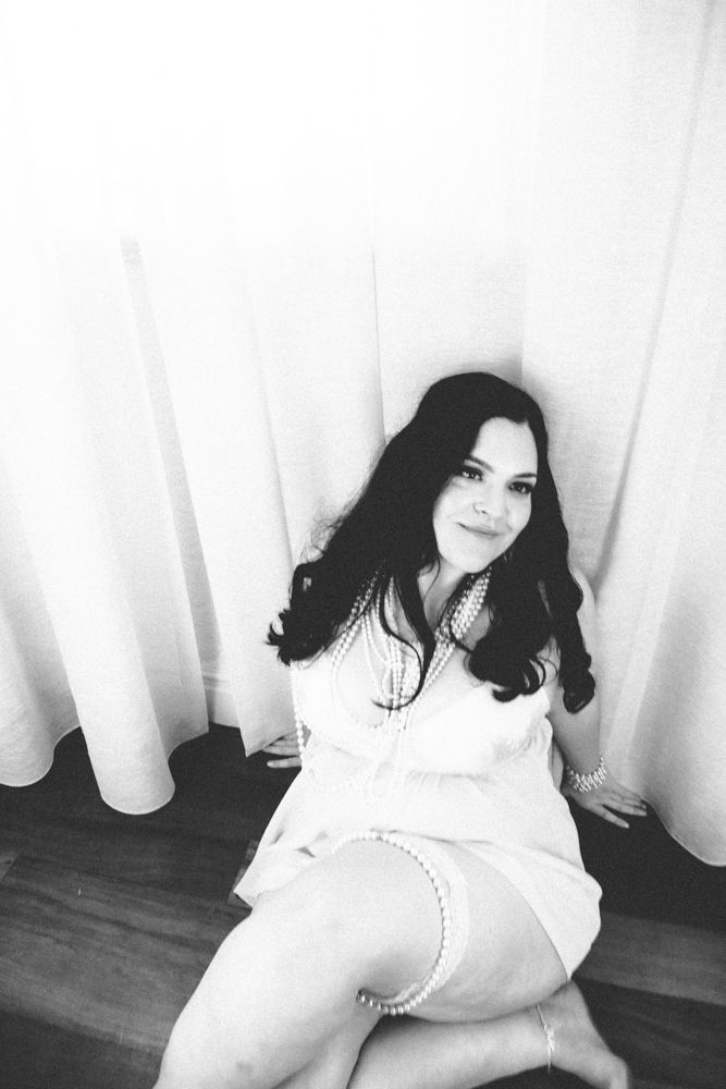 Black and white portrait in casual pose on hardwood floor with white curtain backdrop.