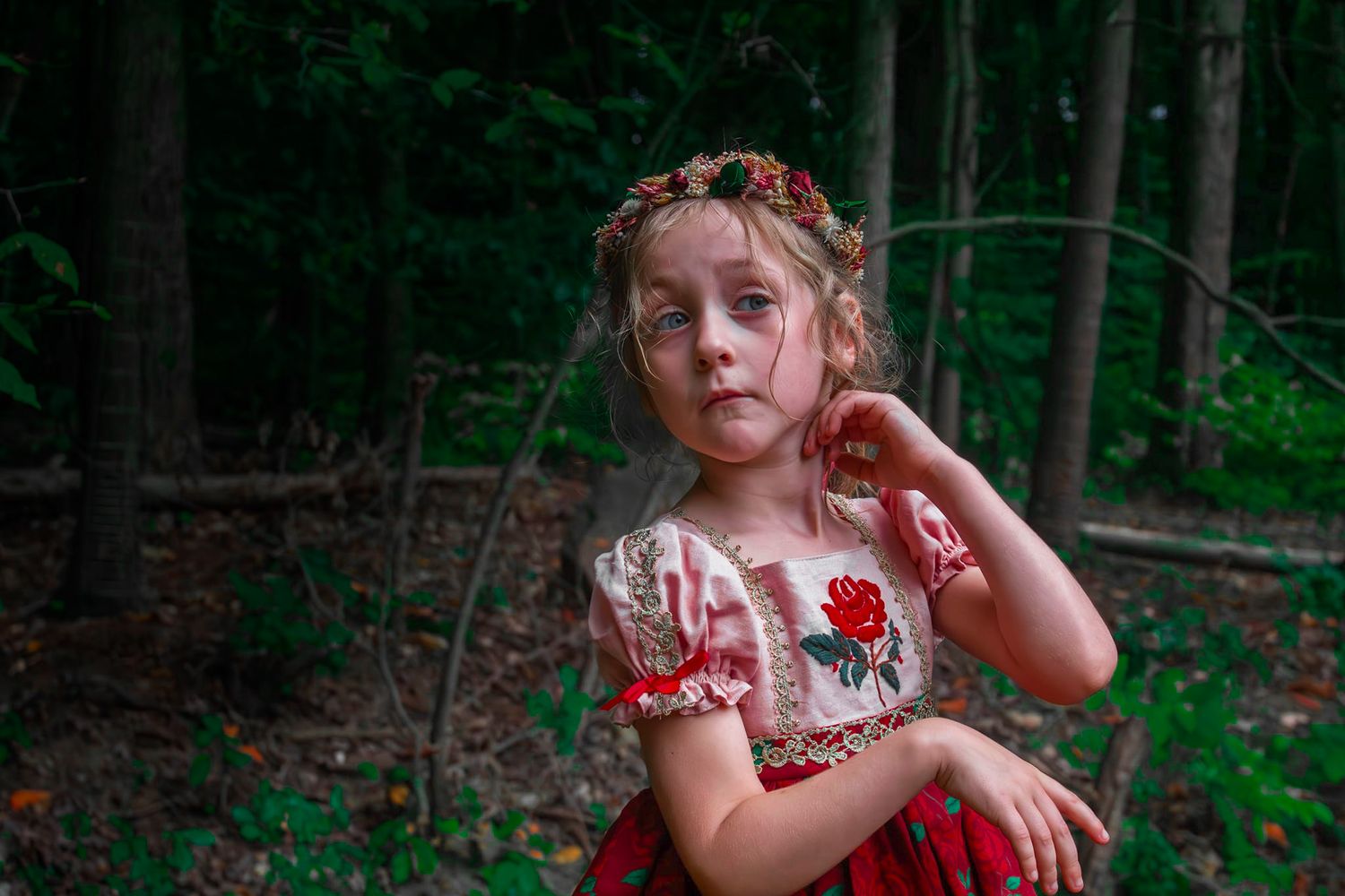Young girl in embroidered rose dress with floral crown standing in a lush green forest setting.