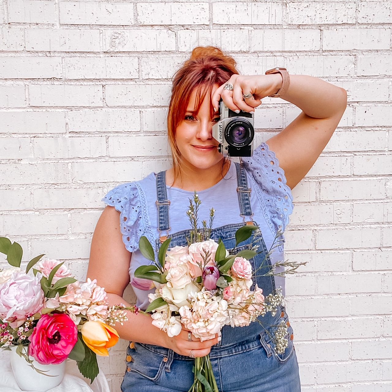Red-haired woman in overalls holding camera and flowers against white brick wall background.