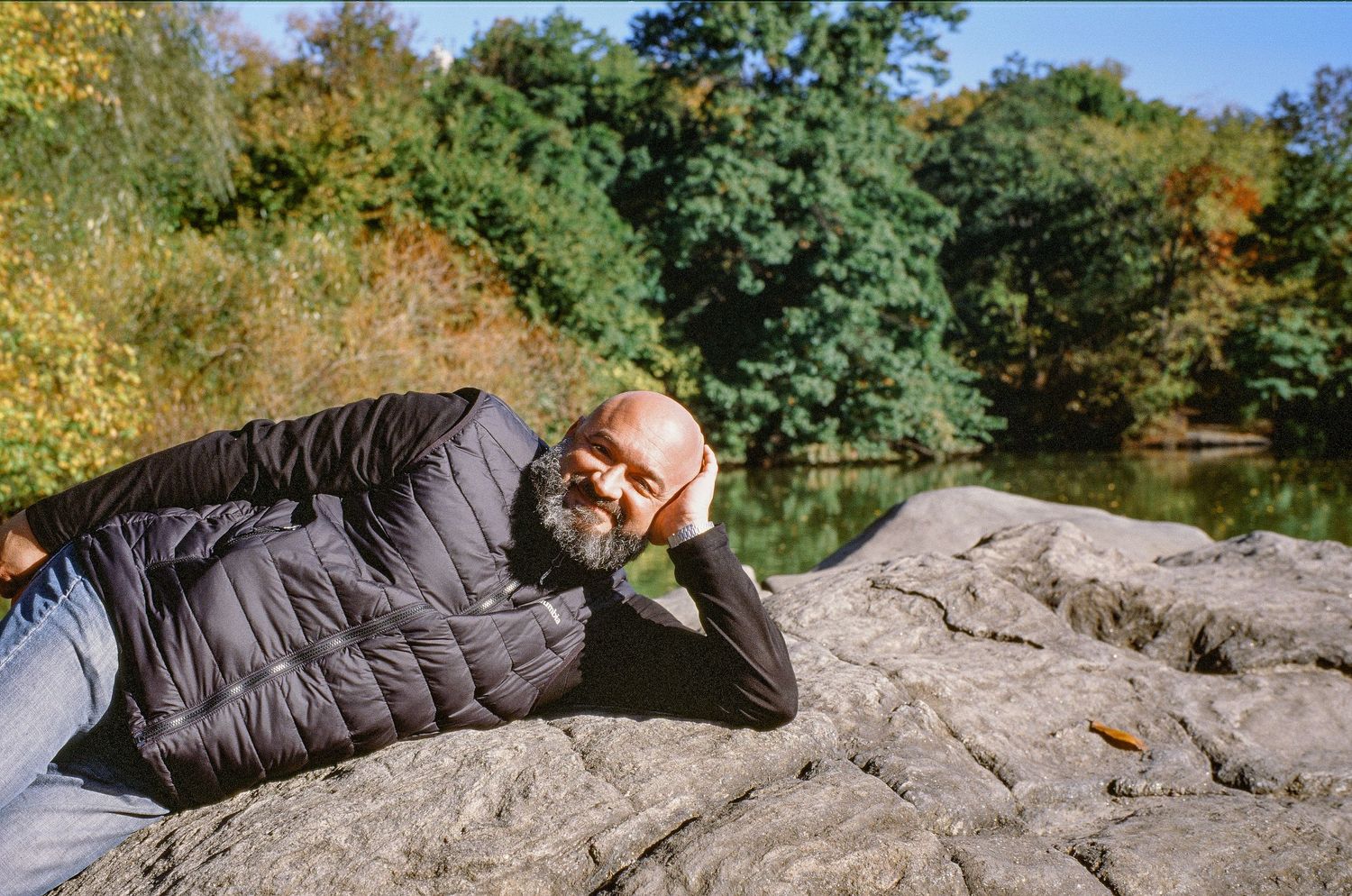 Person relaxing on large rocks near a pond surrounded by autumn trees on a sunny day.