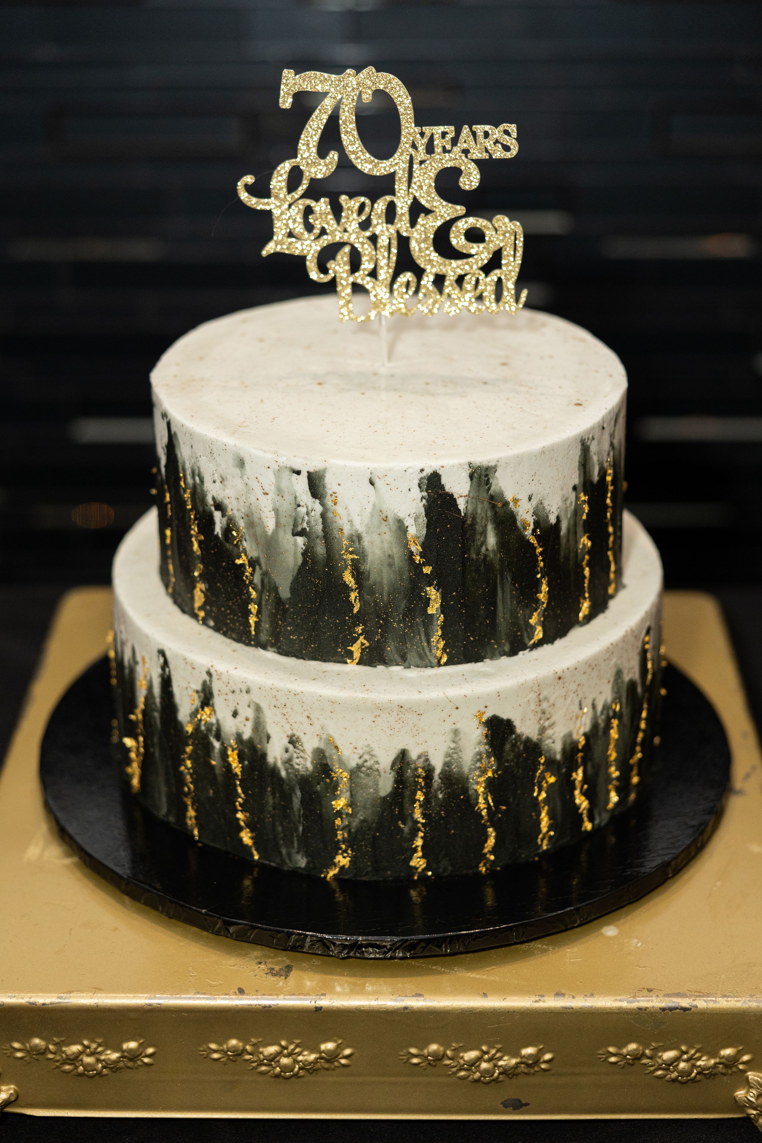 Two-tier black and white cake with gold drip effect and 70th anniversary topper.