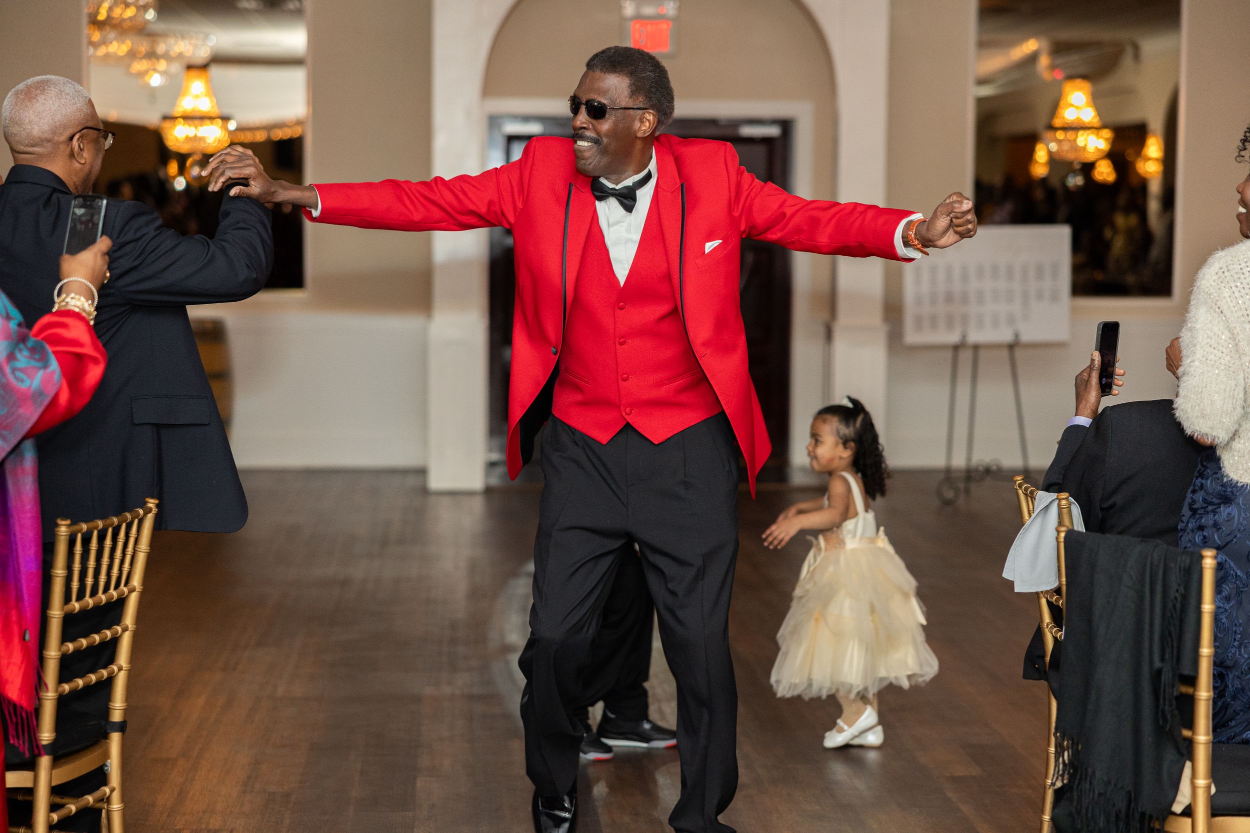Person in red blazer and black pants dancing with arms spread wide at an elegant venue.