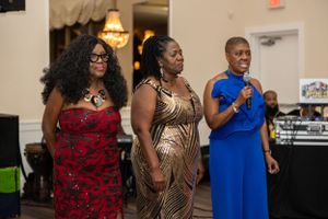 Three women in formal attire speaking at an event with a DJ booth visible in background.