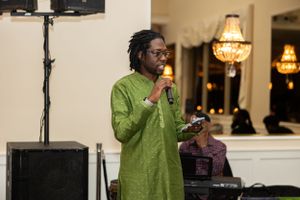 Speaker in green traditional attire addresses audience at formal event with chandelier lighting.