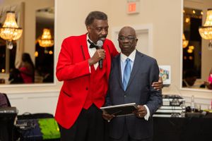 Two people in formal attire presenting at event with DJ equipment in background.