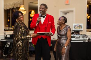 Three people in formal wear interacting near DJ booth at elegant venue with chandelier.