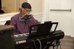 Musician playing Yamaha keyboard at formal event wearing plaid shirt and cap.
