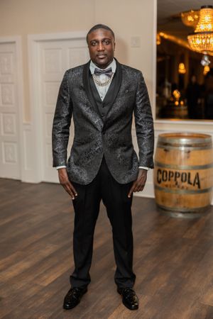 Man wearing a formal gray patterned suit jacket poses in an elegant venue with chandelier and wine barrel.