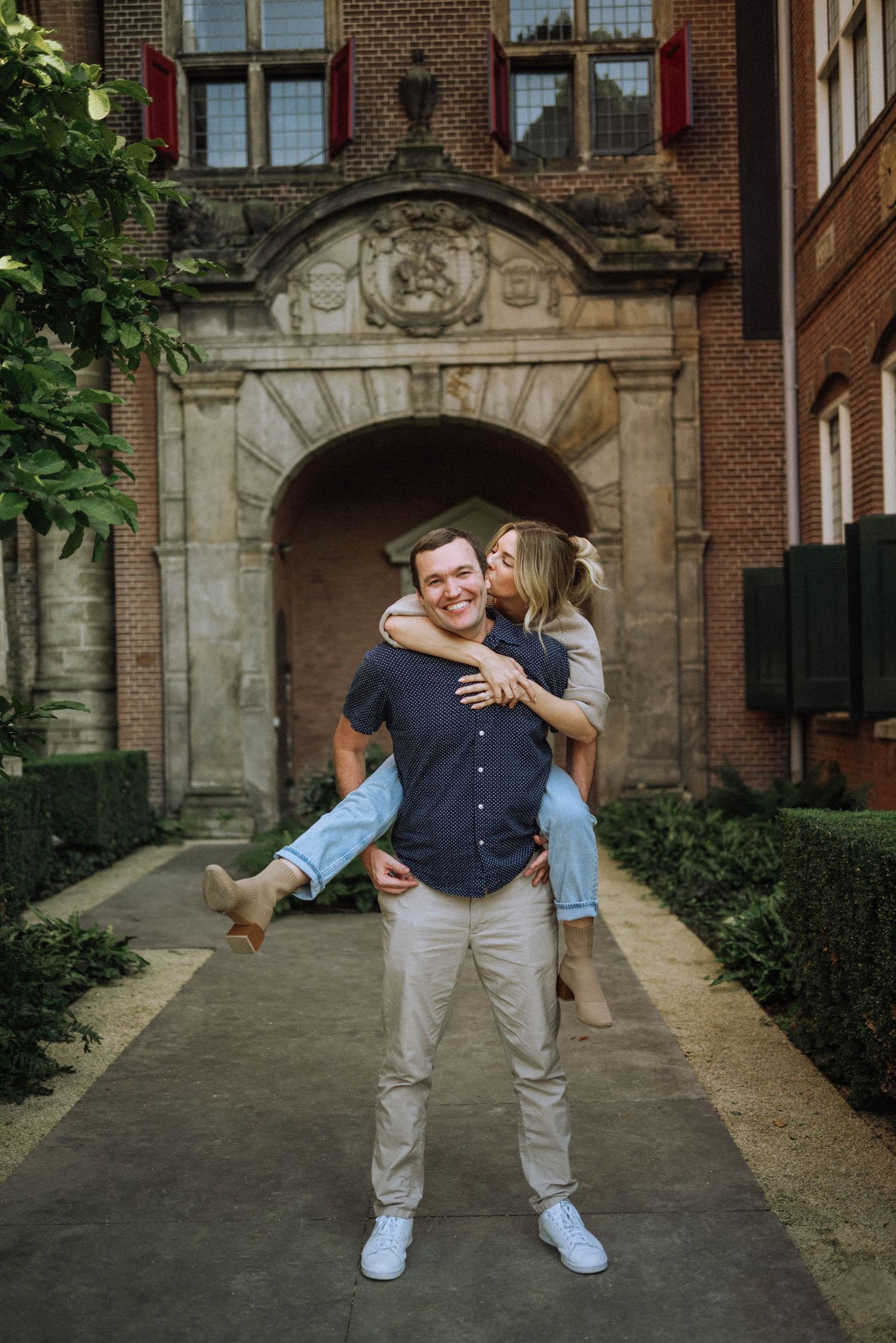 Playful couple in casual, coordinated outfits; woman on man’s back, laughing, in a courtyard with Dutch architecture in the background.