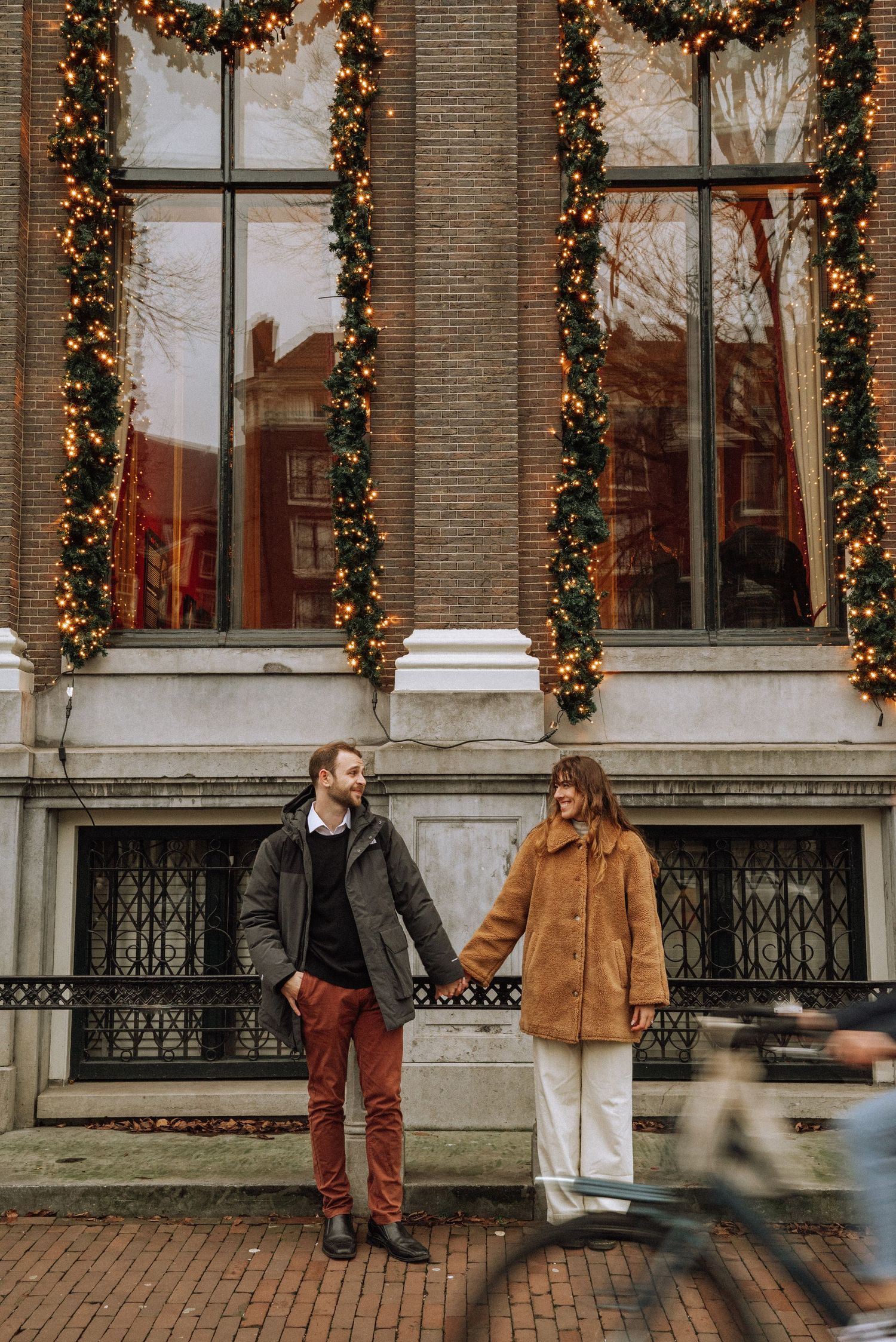 Couple holding hands and laughing in front of a decorated historic Amsterdam building, dressed warmly for a winter proposal photoshoot.