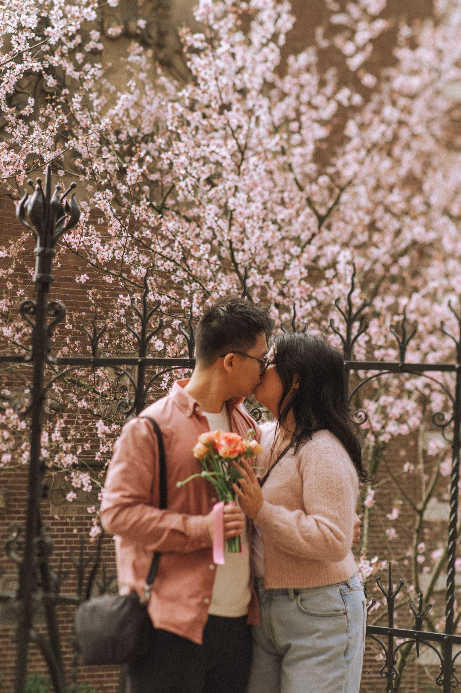 Couple in soft pink and cream outfits sharing a bouquet and a kiss beneath blooming cherry blossoms during a spring Amsterdam proposal