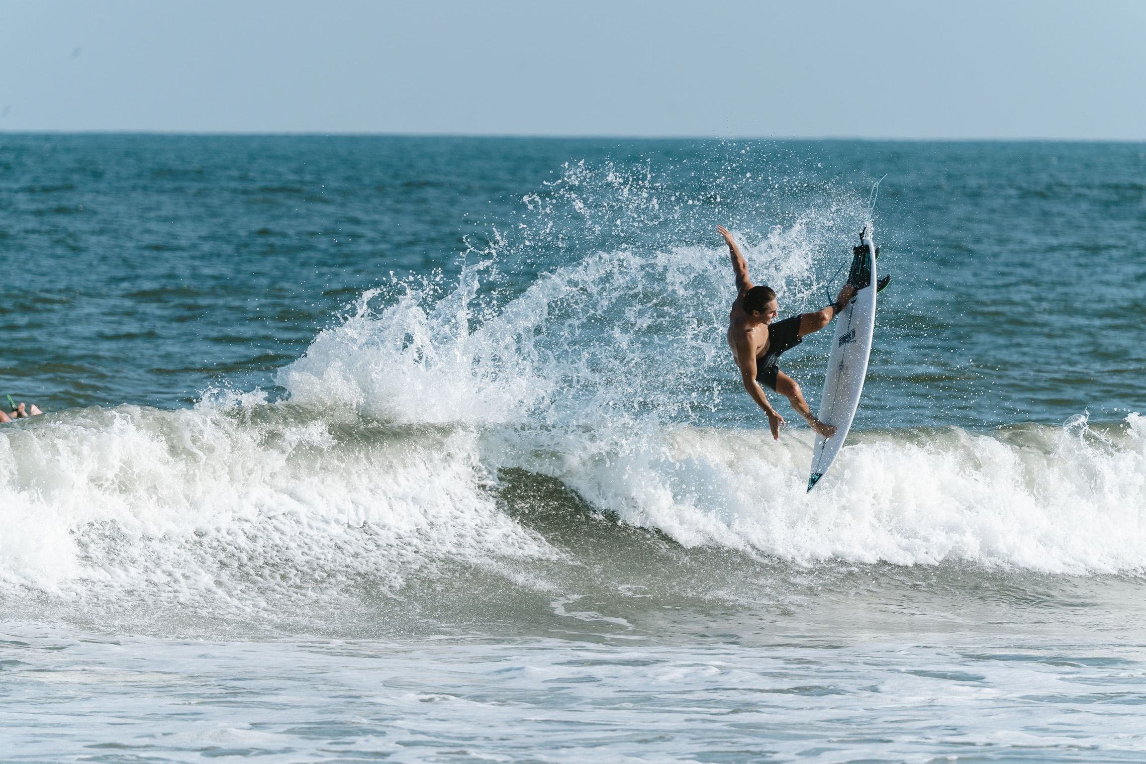Surfer performs aerial maneuver off ocean wave with dramatic splash and spray in bright sunlight.