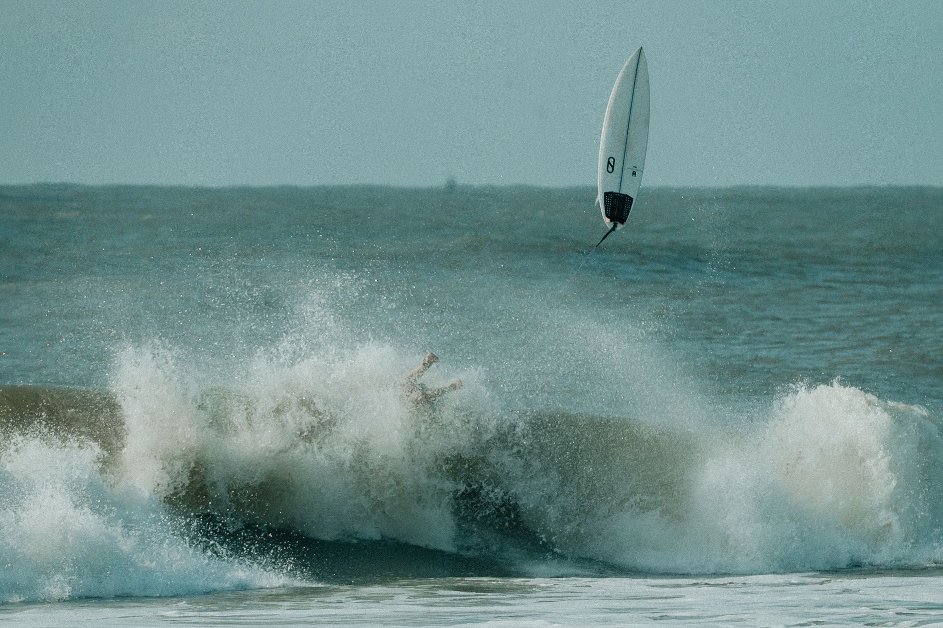 A surfer rides through a powerful ocean wave creating dramatic white spray against a turquoise sea backdrop.