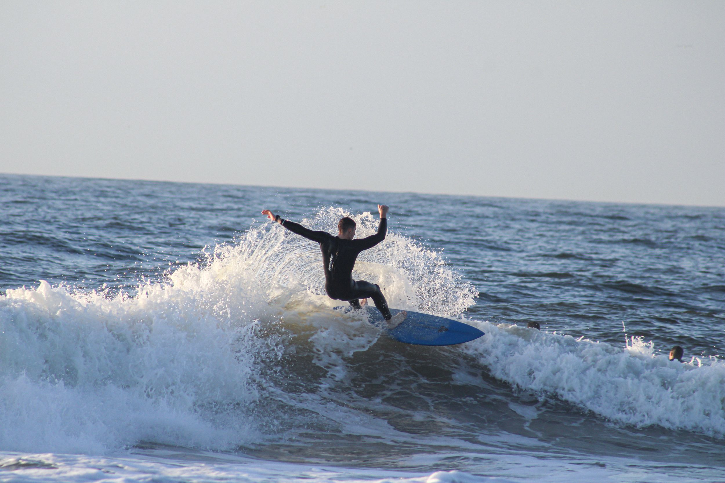 A surfer rides a wave and performs a dynamic maneuver against a gray ocean backdrop.