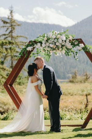 Wedding ceremony under hexagonal arch decorated with white flowers against mountain backdrop.