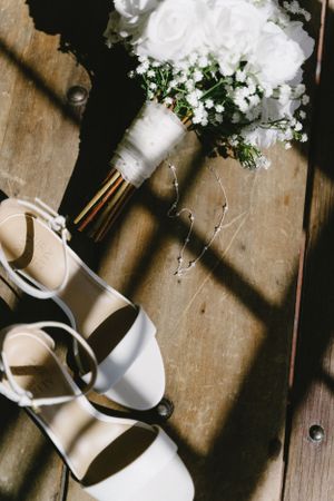 White floral bridal bouquet and elegant ivory heels on rustic wooden floor with dramatic shadows.