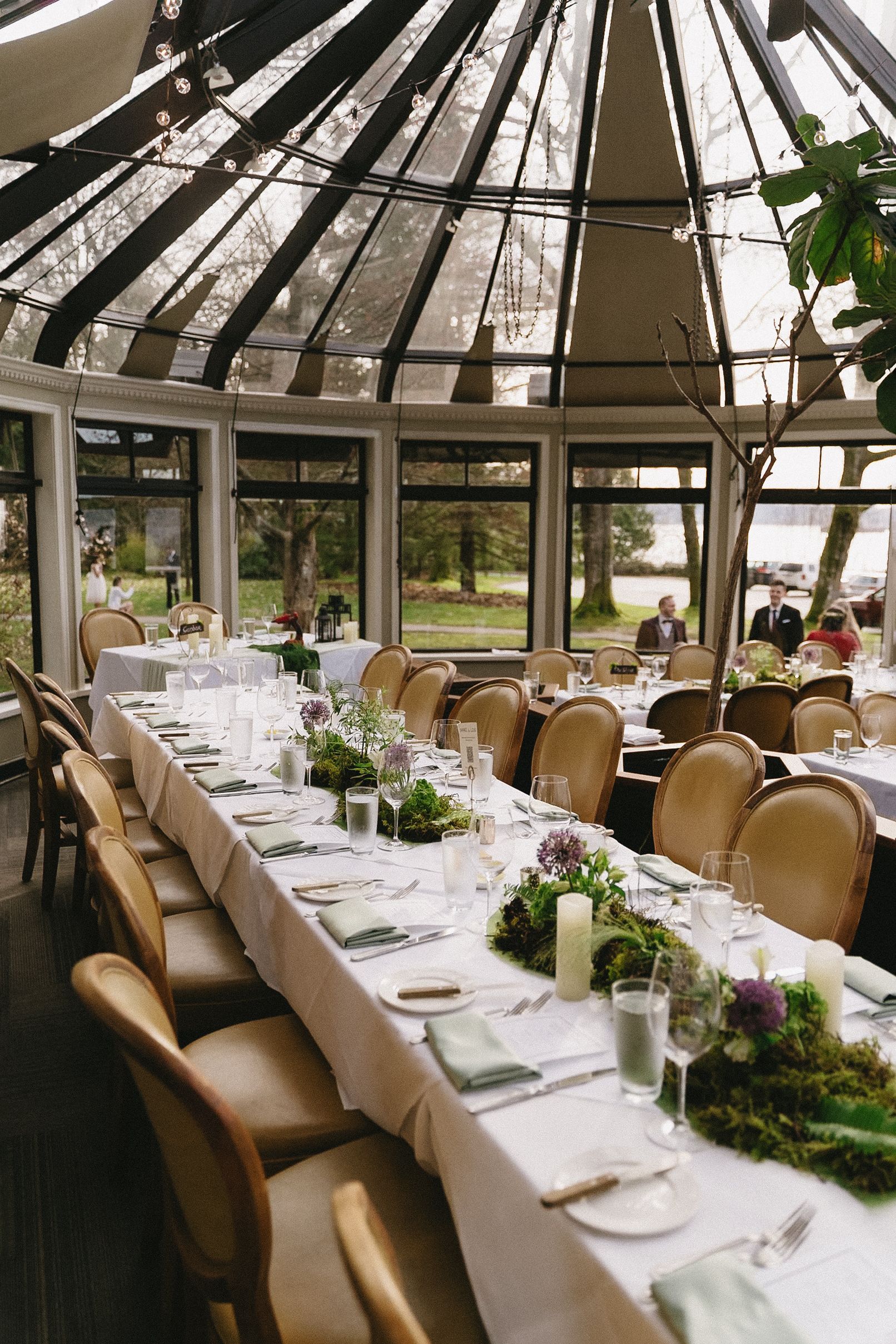 Elegant wedding reception table setting under glass ceiling with greenery runners and gold chairs.