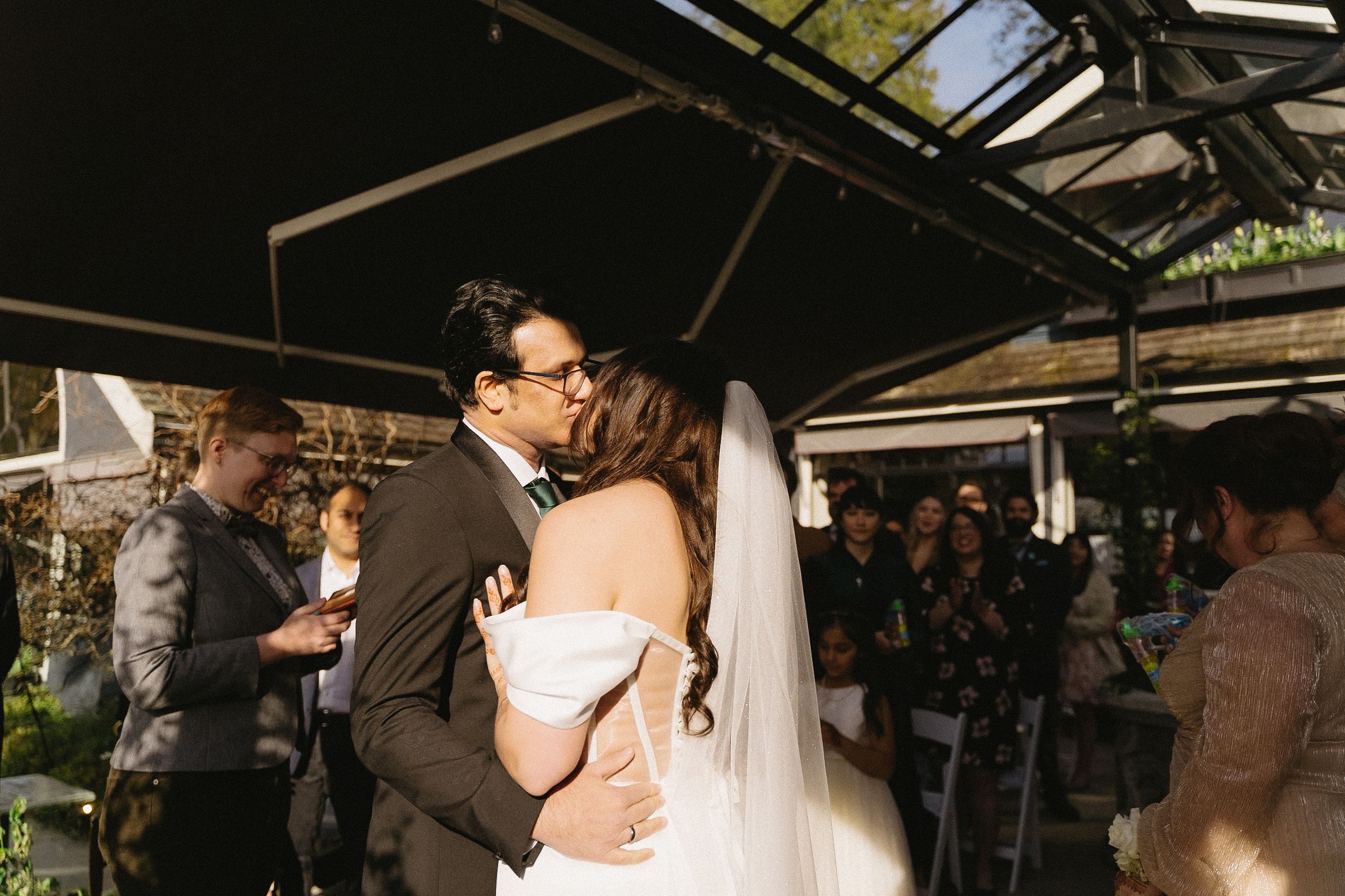 Outdoor wedding ceremony under glass canopy with couple embracing during vows.