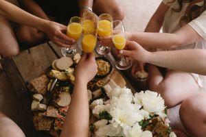 Hands raise mimosa glasses in a toast over a brunch table with white flowers and food.