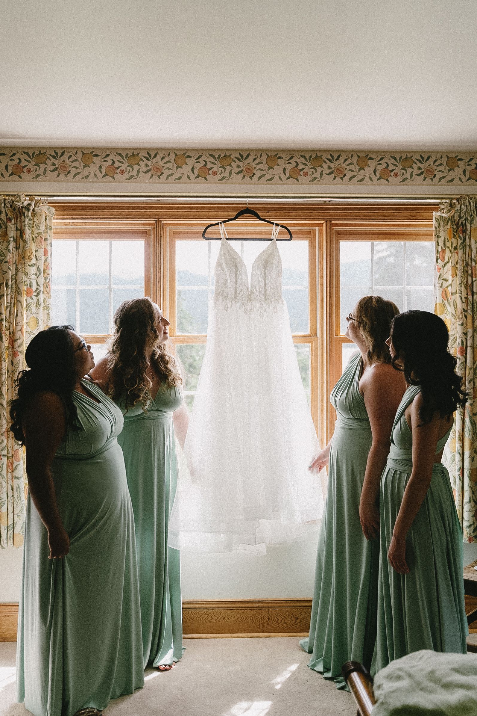 Bridesmaids in mint green dresses admire a white lace wedding gown hanging in front of sunlit windows.